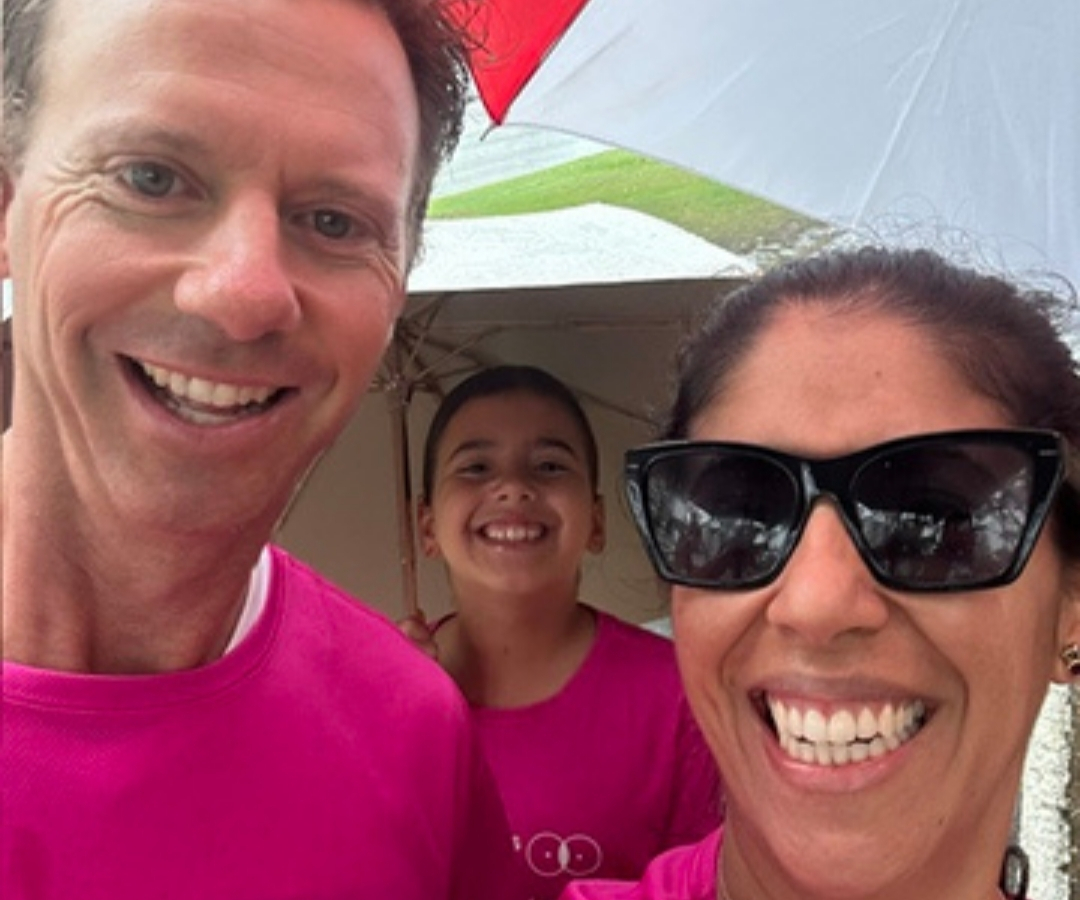 Three people wearing bright pink shirts smile under a red and white umbrella on a cloudy day.